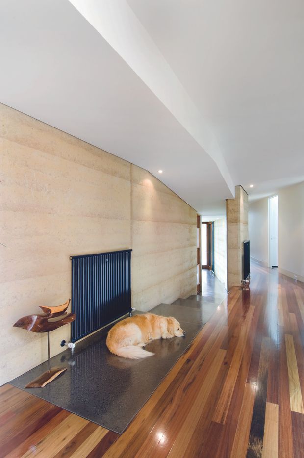 A bright modern residential hallway with a long polished timber floor, warm beige stone or concrete feature walls, a dark vertical-panel radiator, recessed ceiling lights, and a broad angular ceiling bulkhead. A dark floor mat with sculptural wooden decor and a resting dog occupies the foreground, while glazed openings and doorways extend down the corridor.
