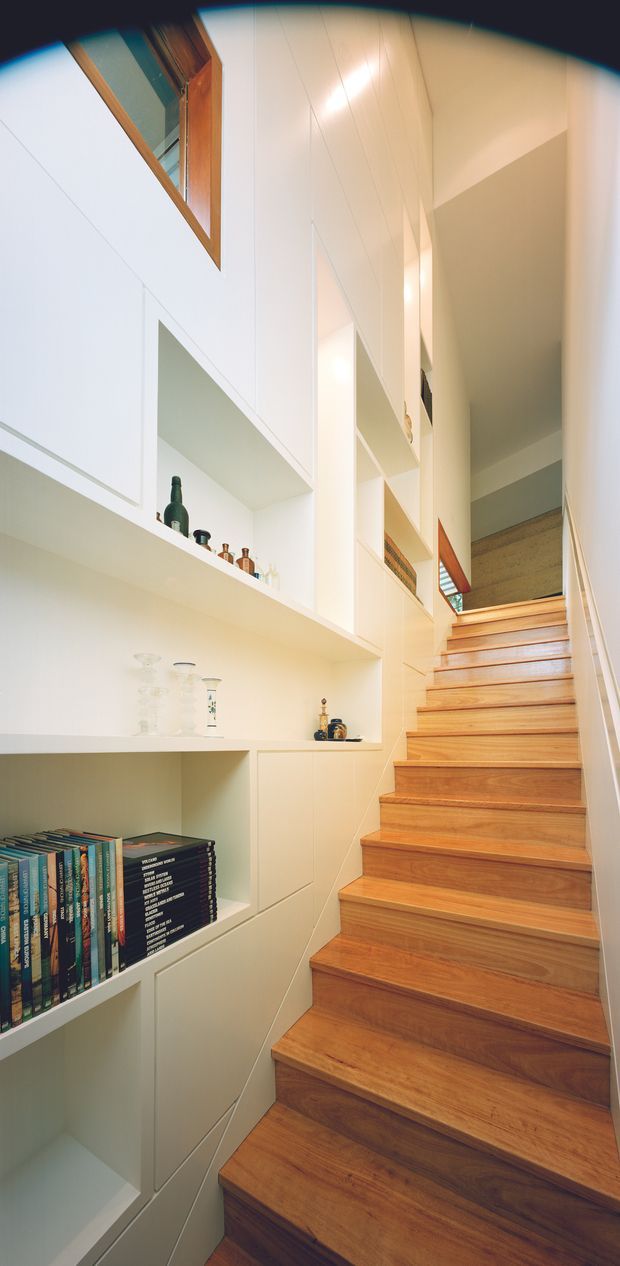 Vertical interior photograph of a bright, narrow stairway rising through a tall multi-level space. Warm timber stairs contrast with white built-in shelving and cabinetry along the left wall, displaying books and small decorative objects. White vertical wall panels and geometric openings create a clean, contemporary built-in composition, with glimpses of upper-level glazing and an open landing.