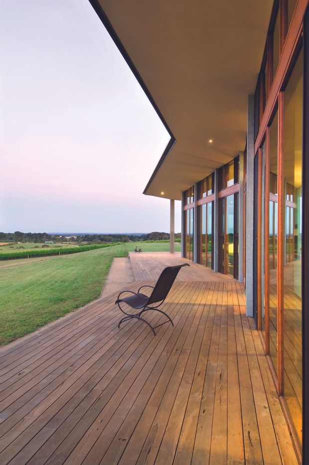A spacious covered timber deck running alongside a contemporary building with extensive floor-to-ceiling glazing and warm timber window and door frames. The deck overlooks an expansive green rural landscape at dusk, with recessed ceiling lights and a single modern lounge chair creating a calm indoor-outdoor setting. The sharply projecting roof canopy and slender supporting columns emphasize the building's horizontal modern form.