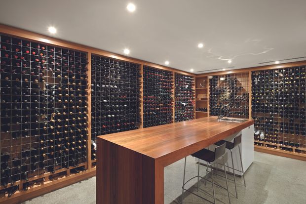 A sophisticated residential wine cellar with extensive floor-to-ceiling bottle storage lining the walls. A large central timber island with a stone or light-coloured counter extension provides a tasting and entertaining area with several bar stools. Recessed ceiling lighting, warm timber framing, dark bottle racks, and polished concrete or stone flooring create a refined, luxurious atmosphere.
