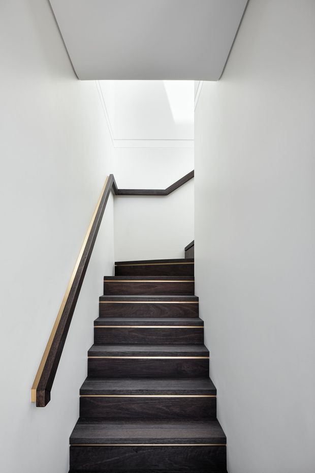 A narrow, minimalist interior stairway with dark timber treads and matching dark timber handrails against smooth white walls. The stairs rise toward a bright upper landing beneath a large skylight or rooflight, creating a clean, high-contrast composition.