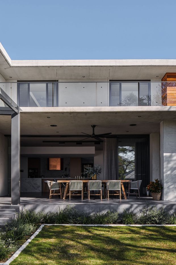 A contemporary two-storey concrete residence viewed from the landscaped front garden. The facade features broad flat roof slabs, exposed concrete soffits and columns, extensive glazing, and a covered outdoor dining terrace with a long timber dining table and chairs. A partially visible kitchen and living area sits behind the terrace, while planted garden beds and lawn occupy the foreground. The design has strong indoor-outdoor connection, clean geometric lines, and a restrained concrete, timber, and glass palette.