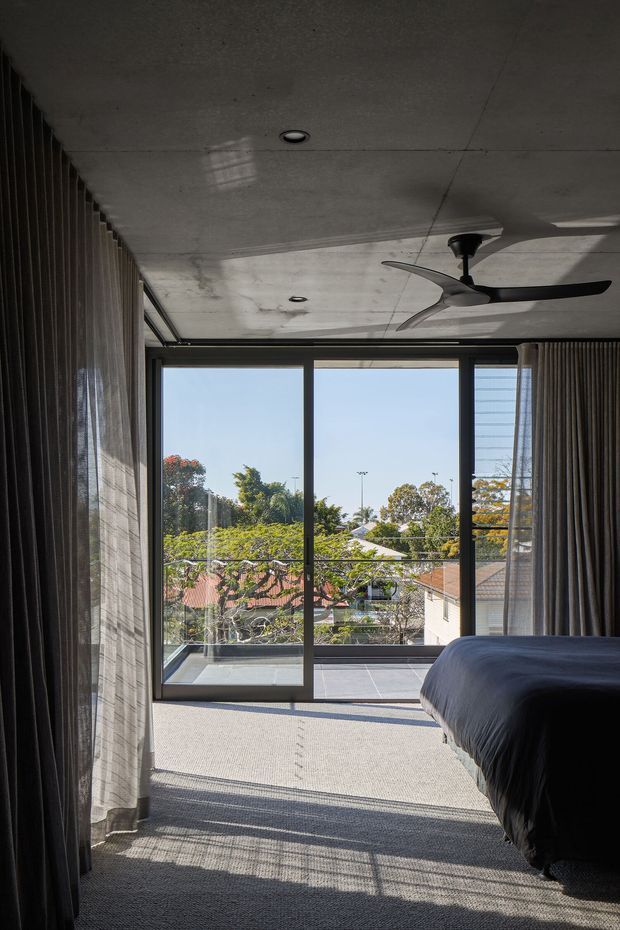 A contemporary bedroom viewed toward a full-height glazed balcony opening. The room features dark textured curtains, a grey carpeted floor, a dark bed with blue-grey bedding, a raw concrete ceiling with recessed downlights, and a modern ceiling fan. The large sliding glass doors open to a balcony overlooking a leafy suburban setting.