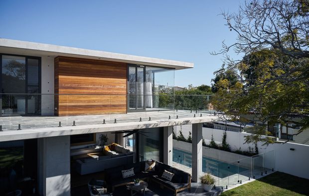 Exterior view of a contemporary multi-level residence with a flat roof, exposed concrete structure, extensive glazing, warm horizontal timber cladding, and glass balustrades. A covered outdoor living area and swimming pool are visible at the lower level, with landscaped surroundings.