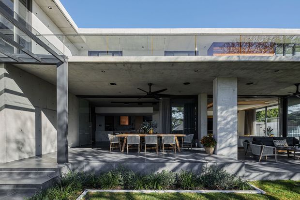 Modern two-storey concrete residence with a broad covered outdoor entertaining area. The image shows a dining setting and lounge furniture beneath a deep cantilevered slab, large glazed openings, exposed concrete columns and soffits, and a flat roofline. Layered garden planting and a lawn sit in front of the terrace.