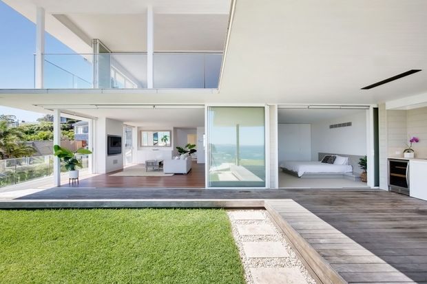 Bright contemporary multi-level home opening onto a landscaped lawn and timber deck. The image shows a white open-plan living area, a bedroom behind sliding glazing, extensive floor-to-ceiling glass, an upper-level void or balcony, and distant ocean views. Clean linear detailing and pale finishes create an airy coastal atmosphere.
