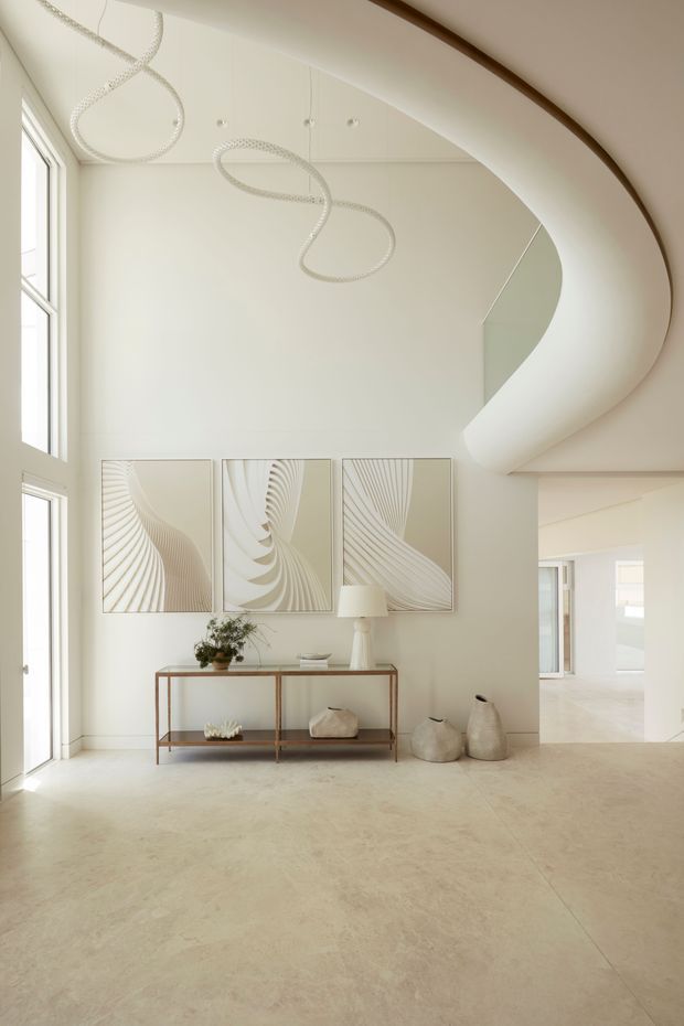 Bright, double-height residential entry hall with a dramatic curved staircase and sculptural loop pendant lights. The space has pale neutral walls, large windows, a light stone or polished concrete floor, a slim console table, abstract artwork and understated decorative objects. An adjacent open circulation area is visible to the right.