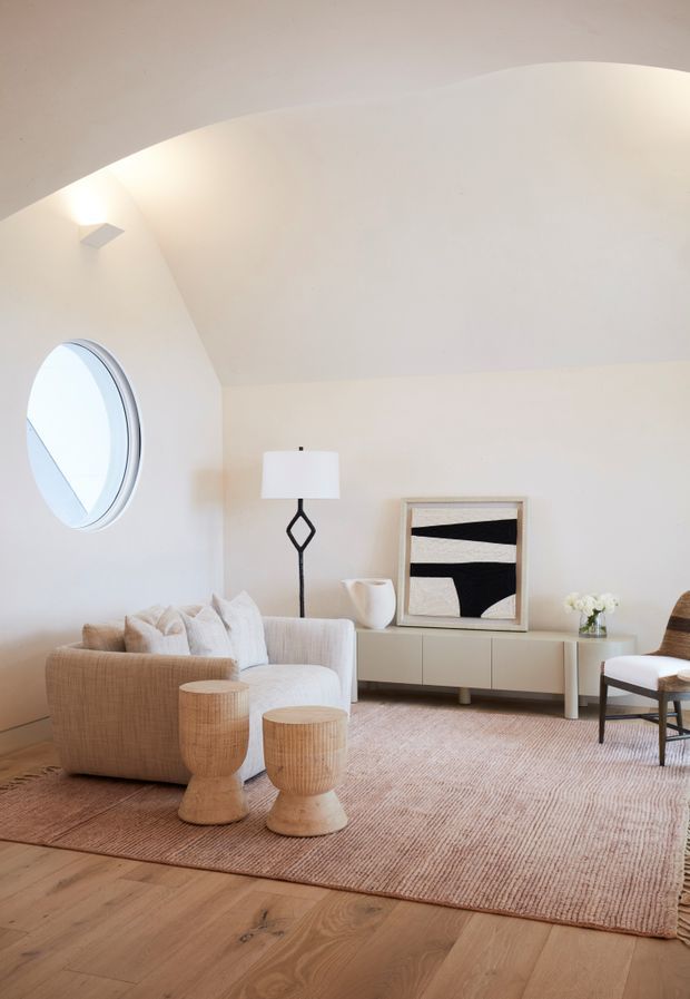 A softly lit, minimalist living room with a sculptural curved white ceiling, round window, pale upholstered sofa, woven wood stools, low contemporary sideboard, geometric floor lamp, framed abstract artwork, and a large neutral textured rug over wide-plank timber flooring.
