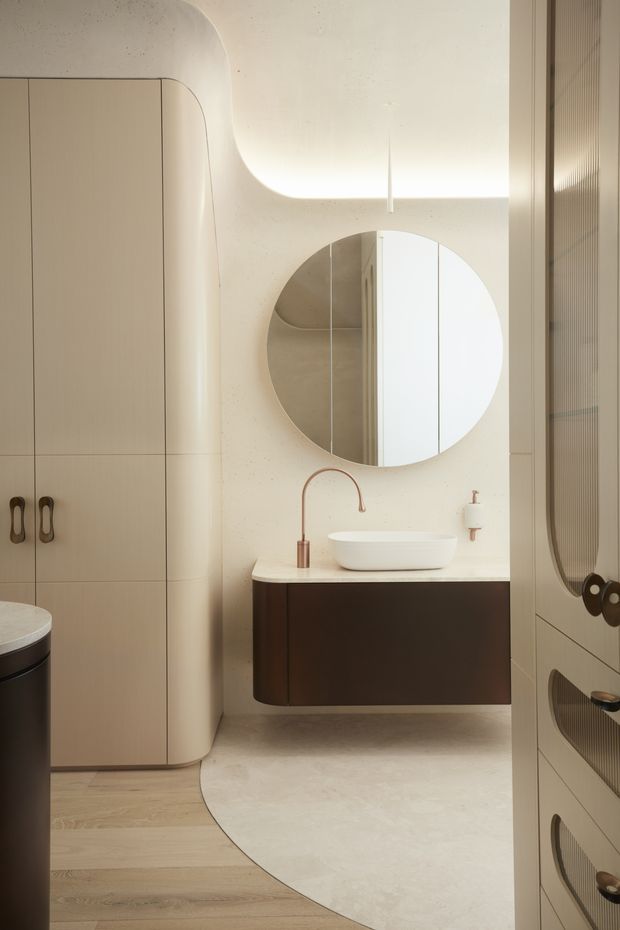 A softly lit contemporary bathroom featuring rounded, sculptural built-in cabinetry, a dark floating vanity with a white vessel basin, a circular wall mirror, and warm neutral finishes. Curved transitions and integrated lighting create a refined, minimalist atmosphere.