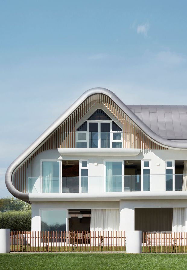 Exterior view of a distinctive multi-storey residence with a flowing, wave-like pitched roof and rounded roof edges. The white facade includes extensive glazing, glass balcony balustrades, vertical timber-toned screening, and curved structural forms. A landscaped lawn and low fence sit in the foreground under a clear sky.