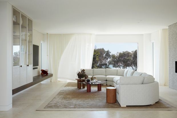 Bright, refined contemporary living room with a large curved sectional sofa arranged around a cluster of small cylindrical coffee tables. Floor-to-ceiling sheer curtains frame a broad window overlooking trees, while full-height built-in cabinetry with open shelving and concealed storage lines the left wall. Pale stone or concrete-look flooring, a neutral textured rug, and a light textured feature wall create an airy, understated interior.
