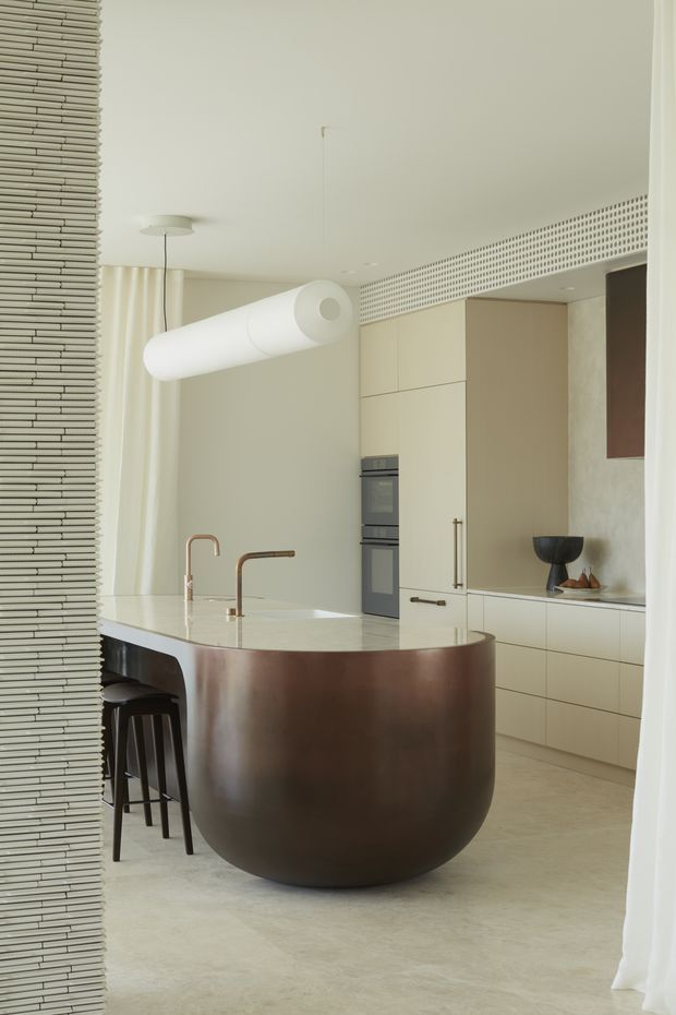 Refined contemporary kitchen featuring a large rounded island with a dark brown curved base and pale stone benchtop, integrated cream cabinetry, built-in ovens, sculptural pendant lighting, and a perforated architectural screen detail near the ceiling. A textured vertical partition frames the left side, while light curtains and a restrained neutral palette create a soft, elegant atmosphere.