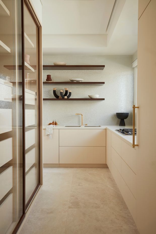 A compact, refined kitchen viewed through a glazed timber-framed doorway. The room features pale integrated cabinetry, a light stone or concrete-look floor, a subtle small-format tiled backsplash, brass-toned tapware and hardware, and dark timber floating shelves displaying ceramics. A cooktop and work surfaces are arranged along the right wall, with soft natural light creating a warm, calm atmosphere.