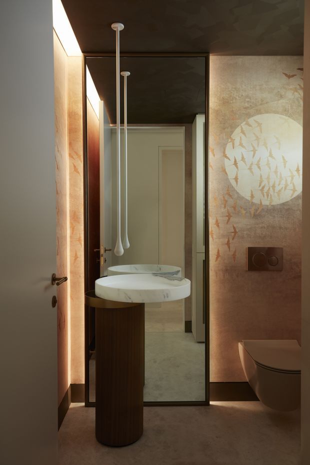 A moody, refined bathroom interior viewed through a partially open door. The space features a sculptural freestanding vessel basin on a dark timber pedestal, a large framed mirror, pendant lighting, a concealed wall-mounted toilet, warm stone or plaster finishes, and a distinctive bird-patterned feature wall with a circular motif. Integrated low-level lighting enhances the intimate atmosphere.