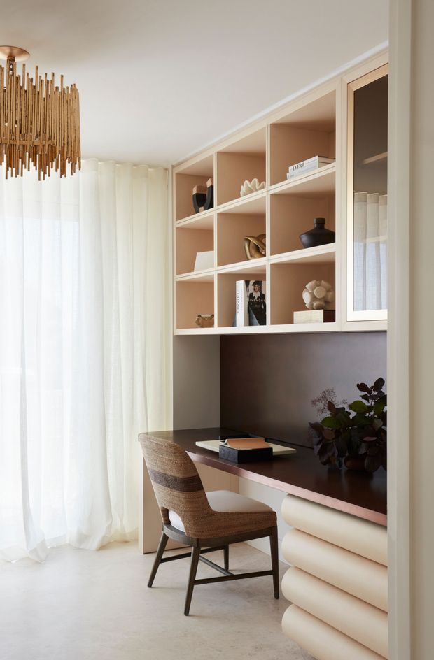 A refined built-in home office nook with a dark desktop, pale peach-beige open shelving, and sculptural rounded drawer fronts below. A woven upholstered chair sits at the desk, while sheer curtains diffuse daylight from a nearby window. Decorative objects, books, and a plant style the shelving, and a linear wood-toned pendant adds warmth.