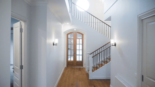 Bright, refined residential hallway and stairway with pale blue-white walls, decorative ceiling cornices, warm timber flooring, and a traditional timber-framed glazed entry door. A staircase with white painted stringers, timber handrails, and slender dark metal balusters rises beside the entry, with wall-mounted sconces providing soft illumination.