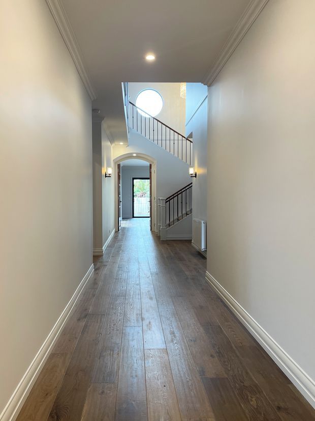 A bright, traditional-style interior hallway with warm timber plank flooring, pale walls, decorative cornices and skirting boards, and a staircase with dark metal balustrades. The corridor leads toward an arched doorway and glazed rear entrance, while a circular window and stairwell bring additional natural light into the space.