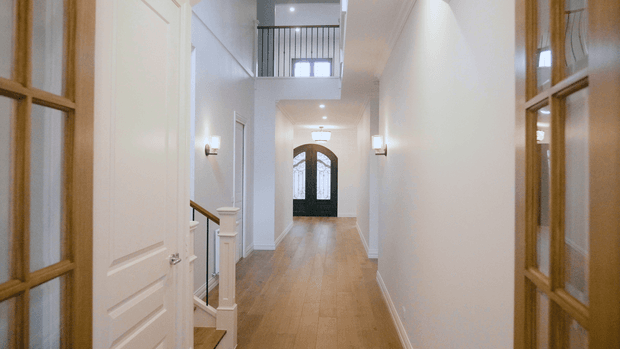 A bright, elegant residential hallway viewed through partially open glazed timber doors. The corridor has warm timber flooring, white paneled walls and doors, wall sconces, a staircase with a timber handrail and white newel post, and a double-height section with an upper-level black balustrade. At the far end is a dark arched glazed entry door.