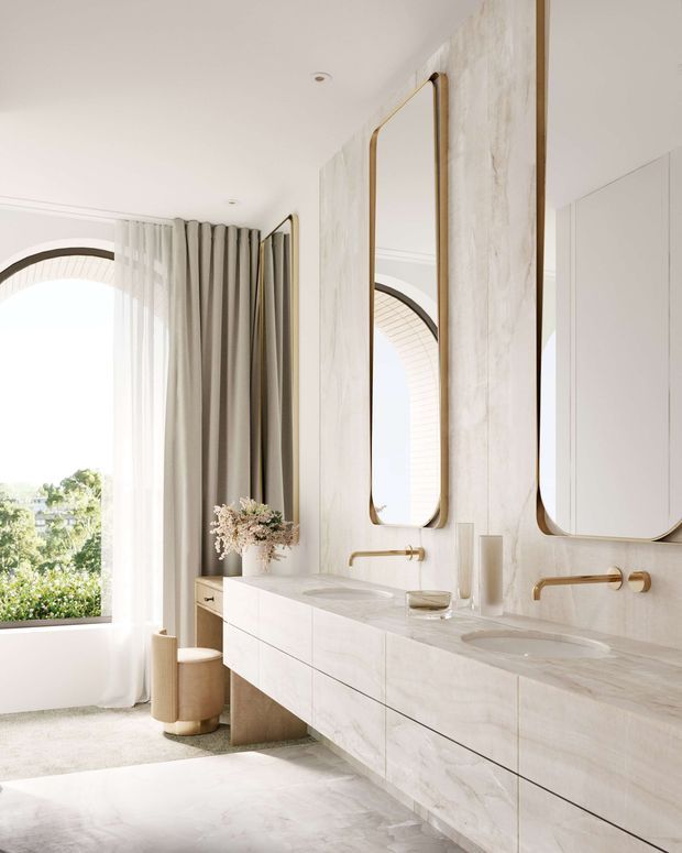 Bright, refined bathroom interior with a long double vanity, two integrated basins, pale stone-look wall and floor finishes, slim brushed-gold wall-mounted tapware, and large rounded-rectangle mirrors. A tall arched window with sheer and taupe curtains brings in abundant natural light, while a small upholstered stool and decorative vase add softness.