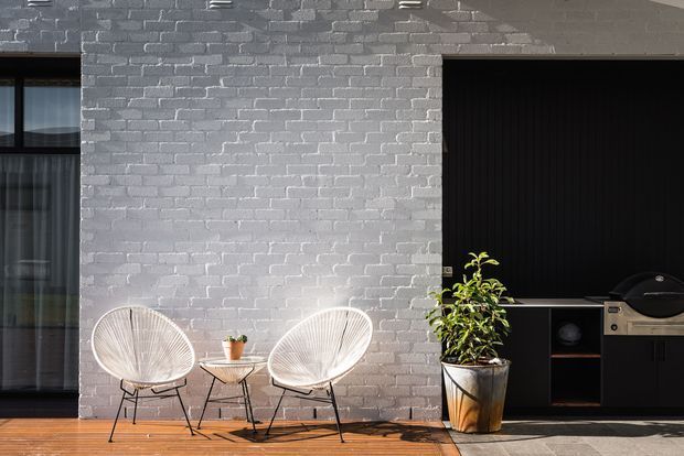 A contemporary outdoor entertaining area beside a modern home, featuring a grey-painted brick wall, dark-framed glazing, timber decking, two white woven lounge chairs, a small side table, potted greenery, and a built-in black outdoor kitchen or barbecue zone under a dark wall opening.