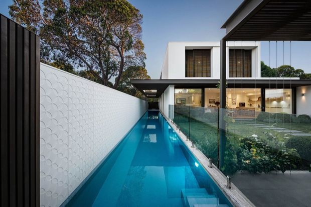 Exterior architectural photograph of a refined modern multi-level residence centered around a narrow linear swimming pool. The composition shows crisp white cubic volumes, extensive floor-to-ceiling glazing, dark metal roof and pergola elements, a transparent glass balustrade, and a patterned white privacy wall running along the pool. A planted courtyard garden and warm-lit interior spaces are visible through the glazing.