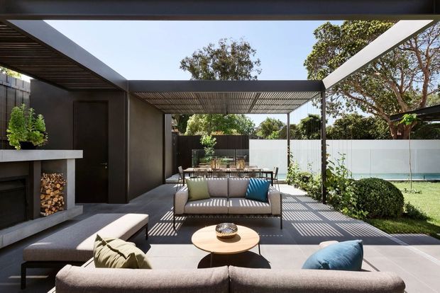 A contemporary outdoor lounge and patio beside a landscaped backyard and swimming pool. The space features a large sectional sofa, armchairs, a round coffee table, a built-in open fireplace with stacked firewood, dark rendered walls, and a slatted pergola creating strong linear shadows. Glazed privacy fencing and mature planting surround the bright, refined entertaining area.