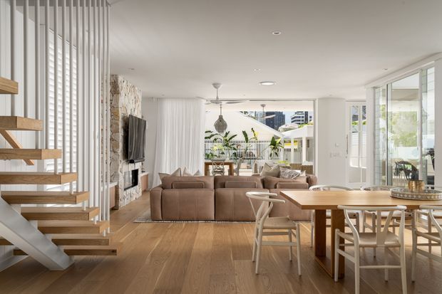Bright open-plan contemporary living and dining area with a timber staircase, warm timber flooring, stone feature wall, large glazed openings, neutral furnishings, and abundant natural light. The living room and dining zone are visually connected, with greenery and exterior structures visible beyond the glazing.