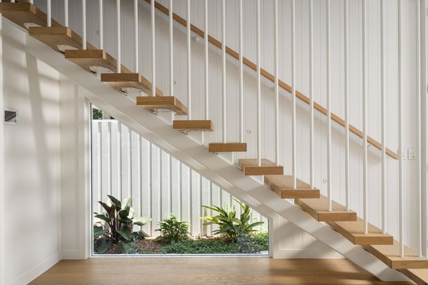 Bright, minimalist stairway interior featuring an open-riser timber staircase with slim vertical white metal rods serving as balustrade, a timber handrail, white vertical wall panelling, and a glazed triangular understair garden with indoor plants. A light timber floor and abundant daylight create an airy, refined atmosphere.