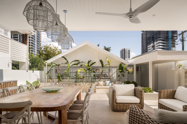 A bright covered outdoor entertaining area with a large timber dining table, woven wicker lounge seating, a ceiling fan, and oversized woven pendant lights. The space opens onto a landscaped courtyard with a white gabled pavilion and climbing greenery, while high-rise buildings are visible beyond. The design has a relaxed resort-like character with neutral upholstery, timber, and natural woven materials.