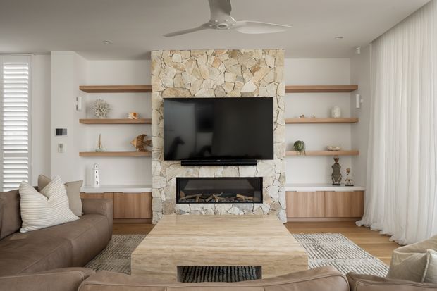 Warm contemporary living room centred on a full-height irregular natural-stone fireplace wall with a built-in television above a linear gas fireplace. Floating timber display shelves and low timber cabinetry flank the fireplace, while neutral upholstered sofas, a large light timber coffee table, layered rugs, sheer curtains, plantation shutters and a ceiling fan complete the room.