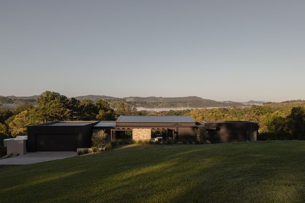 A low, single-storey contemporary residence set into a broad landscaped lawn, with dark horizontal cladding, a shallow corrugated metal roof, an integrated dark garage, and a central light stone section. Mature trees and rolling hills surround the property, with a distant body of water visible beyond.