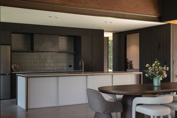 A sophisticated open-plan kitchen and dining area with a dark timber-lined wall, integrated cabinetry, a long pale island with a dark benchtop, and a matching dark oval dining table. Vertical fluted wall detailing, a tiled splashback, warm timber tones, concealed lighting, and sculptural dining chairs create a refined contemporary interior.