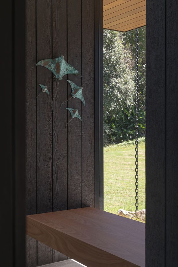 A narrow architectural opening framed by dark vertical timber wall cladding, with a projecting solid timber shelf or bench at the base. A timber-lined soffit and rain chain are visible at the opening, which looks onto a lawn and landscaped greenery. A small decorative metal fish sculpture is mounted on the wall. The image has a refined, dark, natural-material aesthetic with strong contrast between the interior timber and bright garden beyond.