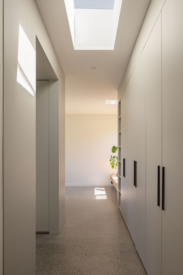 A narrow, bright minimalist interior corridor lined with full-height pale built-in cabinetry and long black vertical handles. A large skylight brings natural light into the hallway, with additional open shelving and a small plant visible at the far end. The floor appears to be a polished or honed terrazzo-like concrete surface, and the overall palette is warm off-white with subtle grey and black accents.