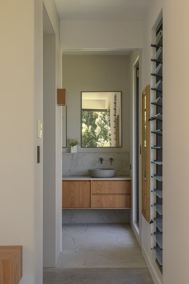 A narrow, refined bathroom viewed from an adjoining hallway, featuring a floating timber vanity with drawers, a round vessel basin, pale marble-look wall and floor finishes, a large rectangular mirror, and dark louvred window shutters. Natural light and greenery are visible through the mirror and side opening.