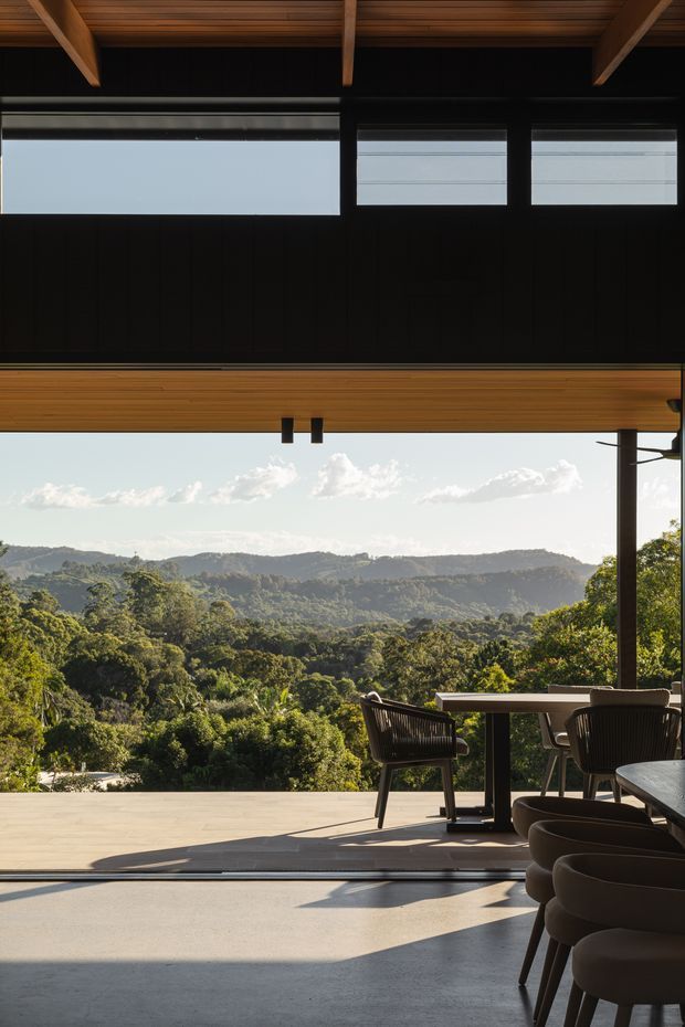 A contemporary covered outdoor dining terrace overlooking a lush forested valley and rolling hills. The space features a broad open glazed frontage, dark vertical wall cladding, warm timber ceiling beams and soffit, a pale paved or tiled terrace, and a dining table with dark woven chairs. The image strongly emphasizes the indoor–outdoor connection and expansive landscape view.