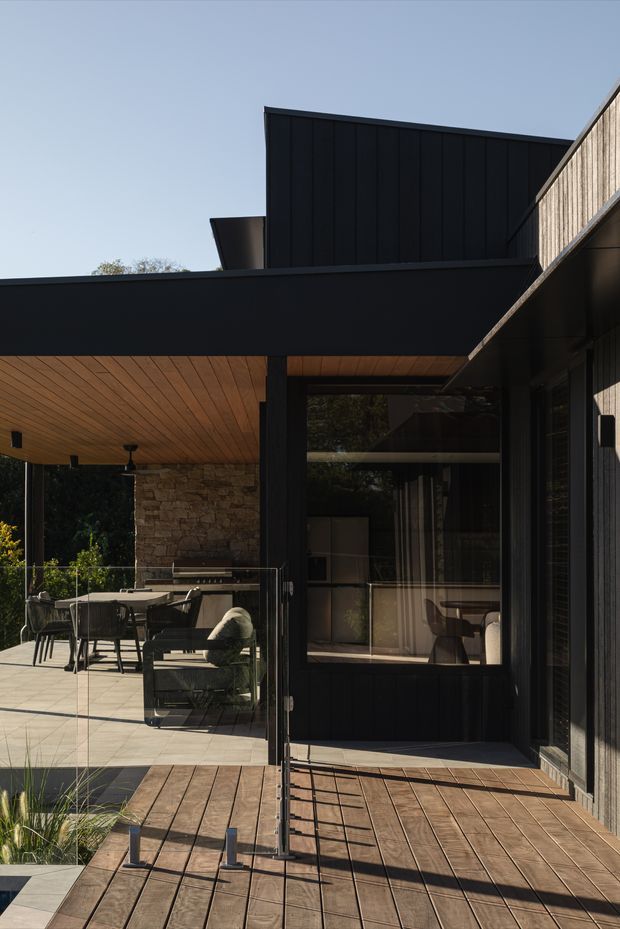 Exterior architectural view of a contemporary dark-clad home opening onto a timber deck and covered outdoor entertaining area. The composition features black vertical cladding, broad projecting roof forms, warm timber soffits and decking, a substantial natural stone wall, full-height glazing, and frameless glass balustrades. Outdoor dining and lounge furniture are visible beneath the covered terrace, with planting around the edges.
