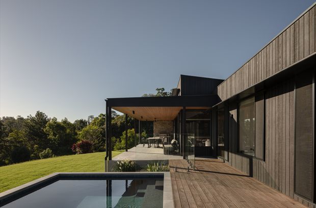 Contemporary low-profile residential exterior viewed from a timber deck, with dark vertical cladding, extensive glazing, a covered outdoor entertaining terrace, landscaped lawn, and a rectangular reflecting or swimming pool in the foreground. The home appears to sit on a leafy elevated site with a strong indoor-outdoor connection and clean geometric lines.