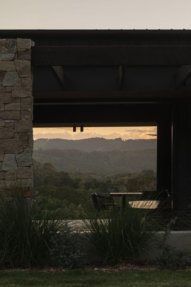 A dark, contemporary architectural opening frames a layered forested valley at sunset. The structure features a substantial rough-stacked stone wall at left, dark timber or metal beams and deep roof overhangs, with built-in or outdoor seating partially obscured by ornamental grasses in the foreground. The composition emphasises the connection between the sheltered building and the surrounding bush landscape.