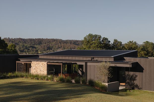 Single-storey contemporary house viewed from the landscaped front yard, with dark vertical cladding, a low-pitched metal roof, extensive glazing, deep covered openings, and a prominent natural stone wall section. The home sits in a leafy, wooded setting with a long horizontal, pavilion-like composition.