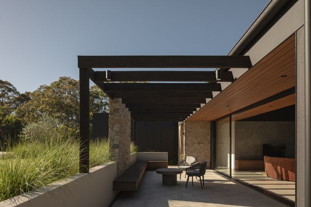 A modern covered outdoor terrace beside a contemporary residence, featuring a dark timber pergola, exposed beams, stone-clad walls, warm timber soffits, large glazed sliding doors, built-in bench seating, and sparse outdoor furniture. Native grasses and trees border the terrace, while the open glazing reveals a refined interior with timber cabinetry.