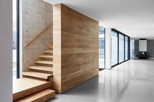 A bright, minimalist contemporary interior centered on a floating timber staircase enclosed by a full-height natural timber feature wall. The stair rises beside a raw exposed concrete wall with a simple timber handrail. Polished light-grey concrete flooring, white structural columns and walls, and expansive black-framed glazing create an open, gallery-like circulation space.