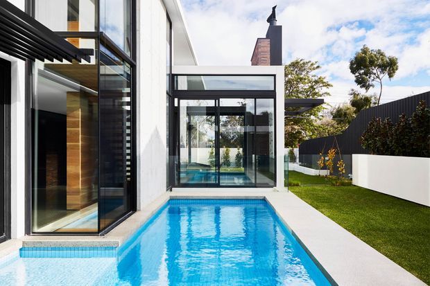 A contemporary residential exterior courtyard viewed alongside a narrow rectangular swimming pool. The scene features crisp white rendered walls, extensive black-framed glazing, a dark metal pergola or louvre element, and a landscaped lawn bordered by white planter walls and dark vertical cladding. The pool extends toward a glazed pavilion or interior space, creating strong indoor-outdoor continuity. Bright daylight, clean geometric lines, and reflective water give the image a fresh, high-contrast appearance.