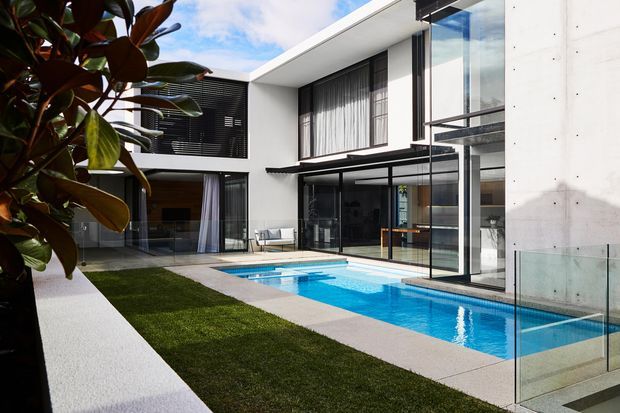 Exterior view of a contemporary multi-level residence arranged around a private courtyard and bright blue swimming pool. The home features crisp white rendered/concrete walls, extensive floor-to-ceiling glazing, dark framed windows, slim black metal awnings, and a glass pool balustrade. A lawn strip and tropical planting soften the geometric architecture.