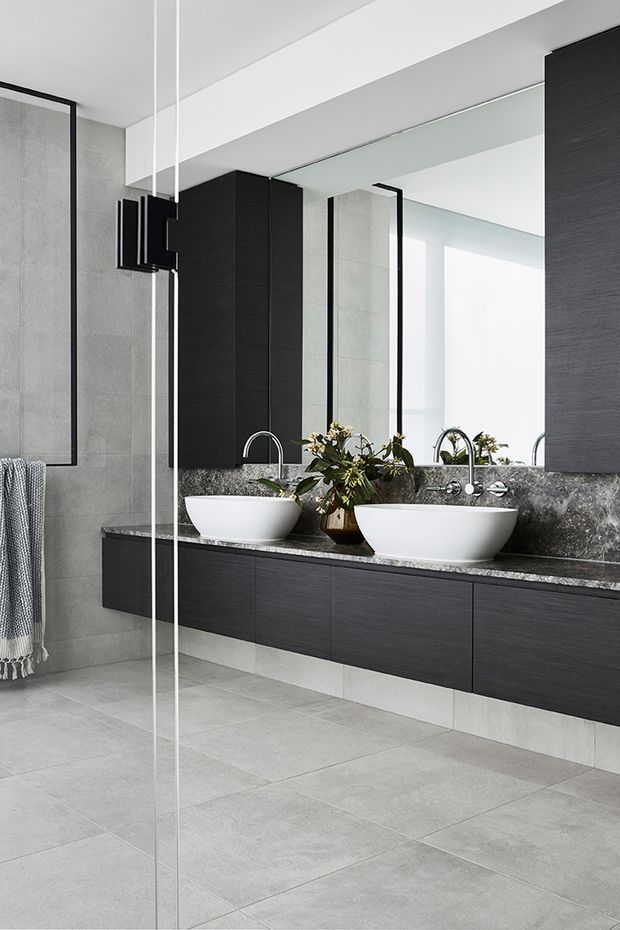 Contemporary bathroom with a double floating vanity, two white vessel basins, dark wood-look cabinetry, a dark natural-stone benchtop, large grey wall and floor tiles, black-framed glass shower screening, and a full-width mirrored wall. The space is clean-lined, monochromatic, and softly lit.