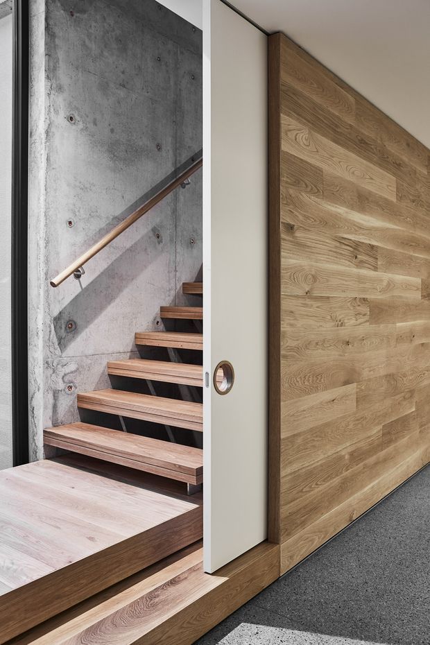 Close-up architectural view of a contemporary stairway and entry transition. Floating timber stairs and a timber platform sit against an exposed concrete wall with a slim metal handrail. A large vertical timber-clad pivot door or wall panel with a circular pull detail dominates the right side, while dark flooring and clean white wall surfaces create a refined modern-industrial composition.