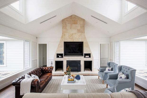 Bright, spacious living room with a vaulted white-paneled ceiling, large windows, a central natural-stone fireplace wall with built-in shelving and television, and a mix of a brown leather sofa, pale blue armchairs, and a light coffee table. The room has a refined, comfortable traditional-coastal character.