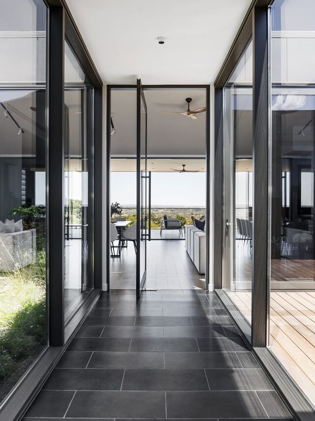 A bright, modern glazed corridor or transitional entry space with dark tiled flooring and extensive black-framed floor-to-ceiling glazing. The corridor opens toward a light-filled living and outdoor entertaining area, with a distant landscape view. Timber decking, contemporary seating, ceiling fans, and strong indoor-outdoor connections are visible beyond the glazing.