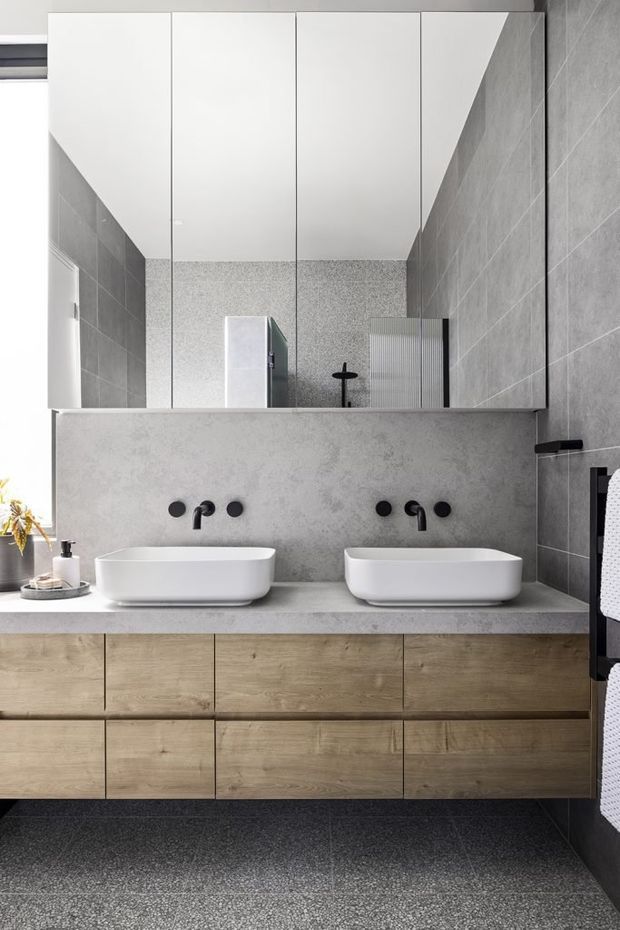 Contemporary double-vanity bathroom with a wall-mounted timber-look vanity, two white vessel basins, matte black wall-mounted taps, and a large mirrored cabinet. Grey tiled walls and a matching grey stone-look benchtop create a restrained monochromatic palette, while the dark speckled floor adds texture.