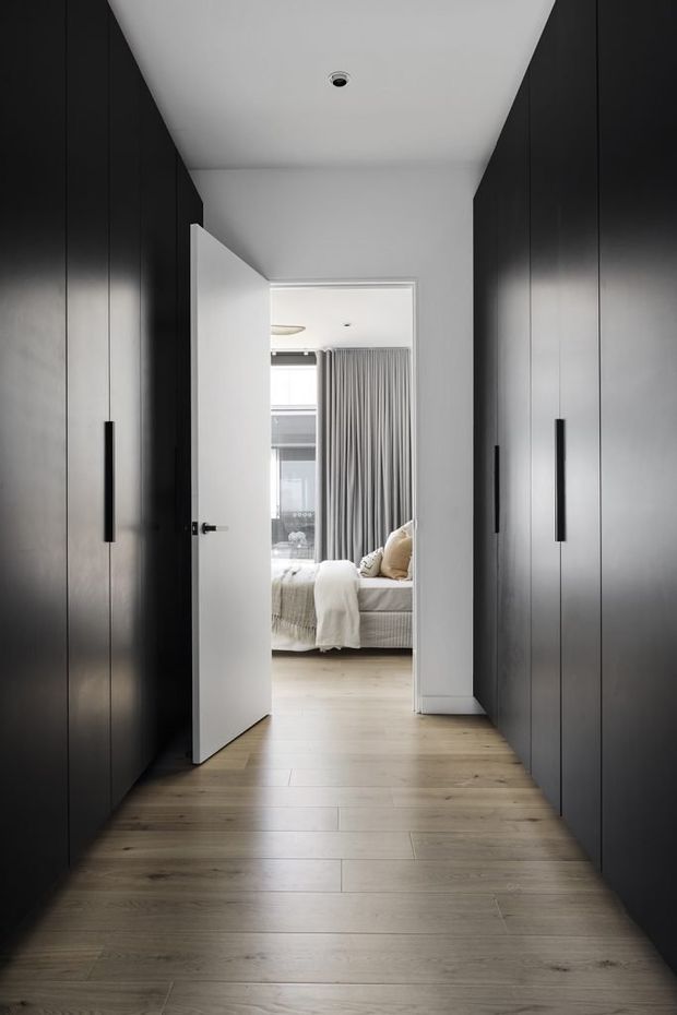 A narrow interior hallway lined on both sides with tall, dark built-in cabinetry featuring long black vertical handles. The hallway has a pale timber-look floor, white ceiling and walls, and a recessed ceiling light. An open white door at the end leads into a softly styled bedroom with a bed, neutral bedding, curtains and a window.